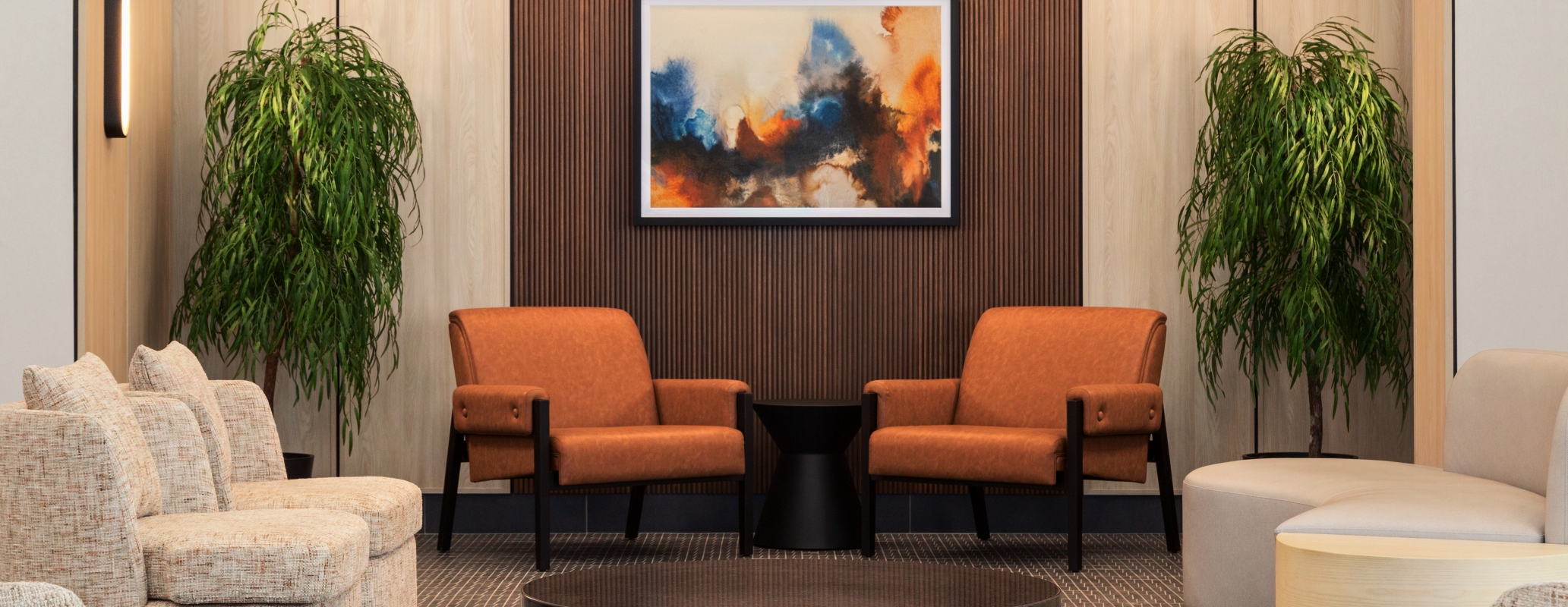 Modern hotel-style lounge with a wood-paneled accent wall, abstract framed painting, orange chairs, and green potted plants.