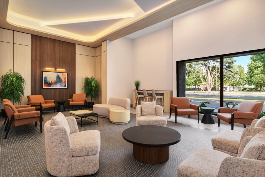 Modern hotel lobby with tan upholstered chairs, round coffee tables, and wood paneled walls near a large windowed wall.