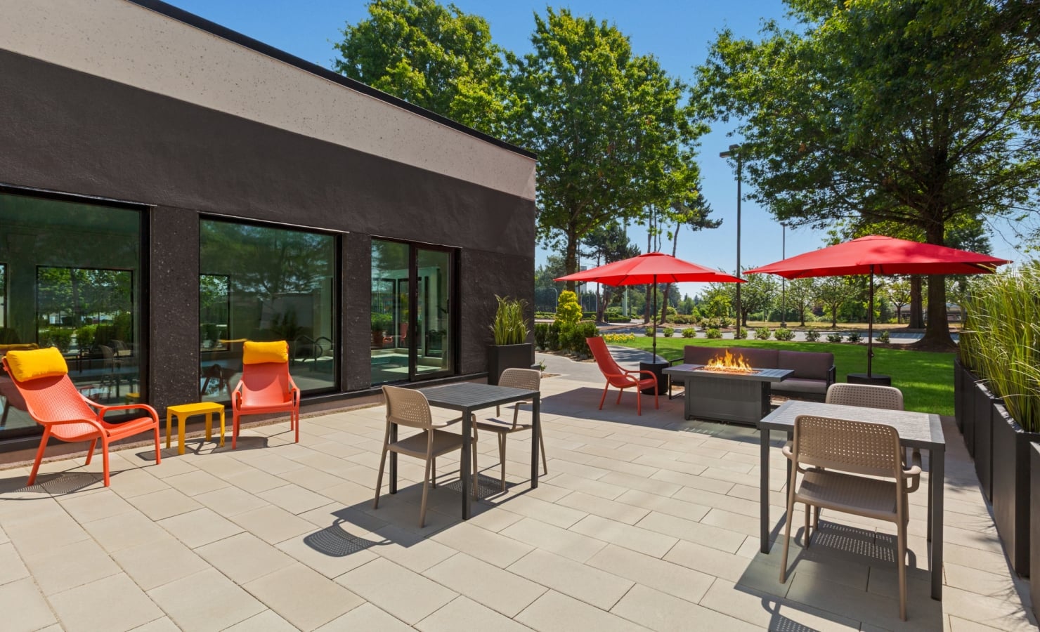 Outdoor patio with a modern dark building, orange chairs, grey tables, red umbrellas, and a fire pit in the seating area.