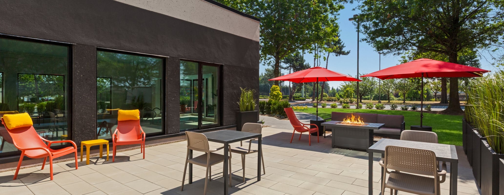 Outdoor patio with a modern dark building, orange chairs, grey tables, red umbrellas, and a fire pit in the seating area.