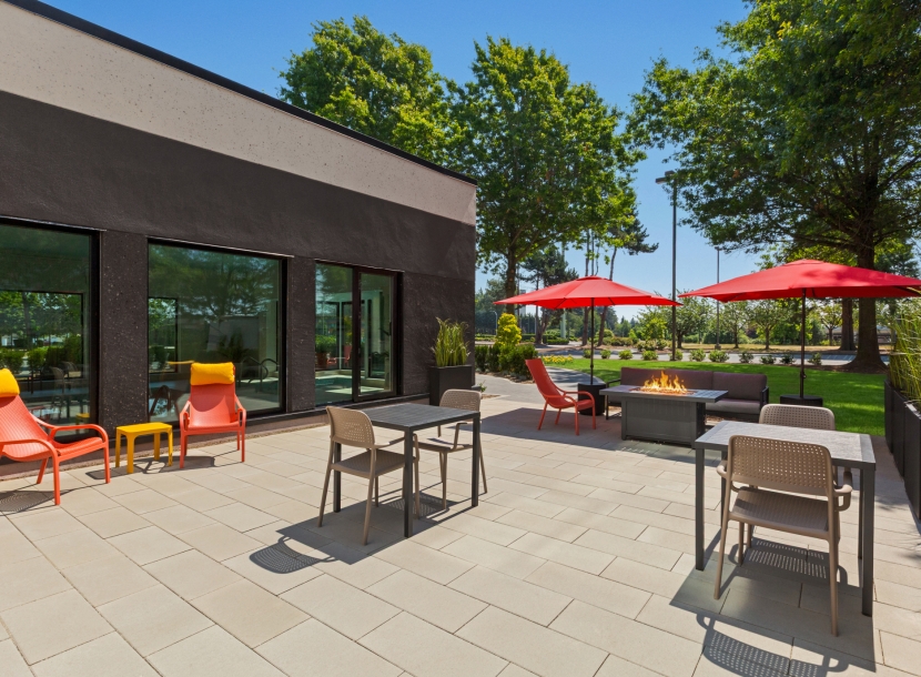 Outdoor patio with a modern dark building, orange chairs, grey tables, red umbrellas, and a fire pit in the seating area.