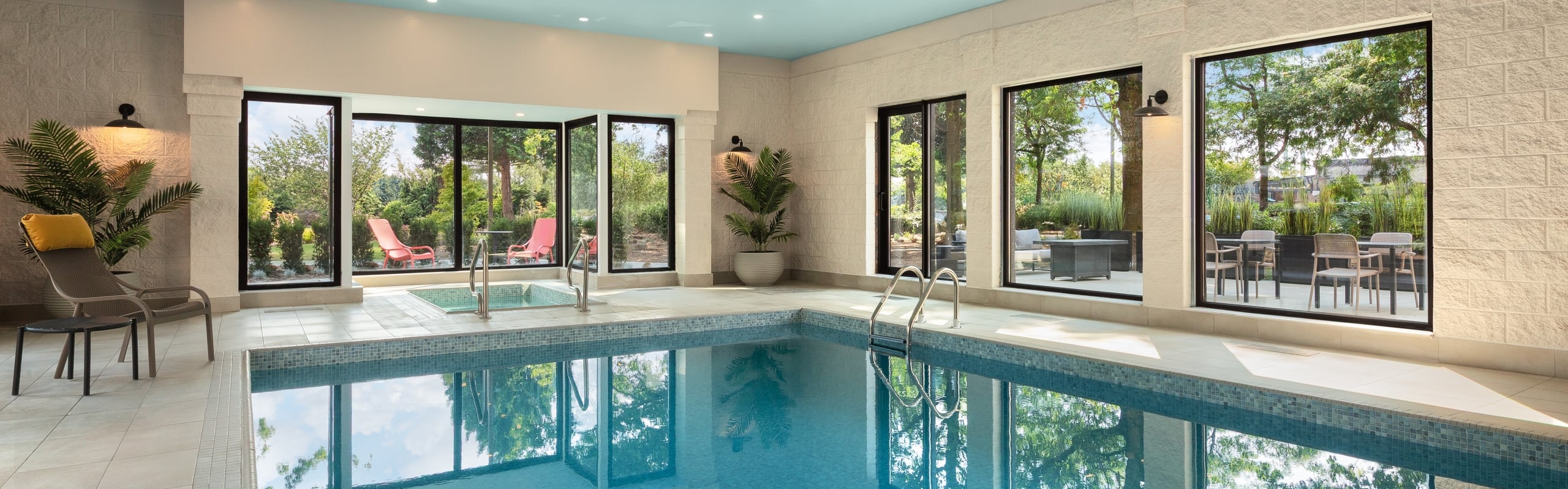 Indoor pool area with a blue ceiling, large windows, and lounge chairs by the poolside plants.