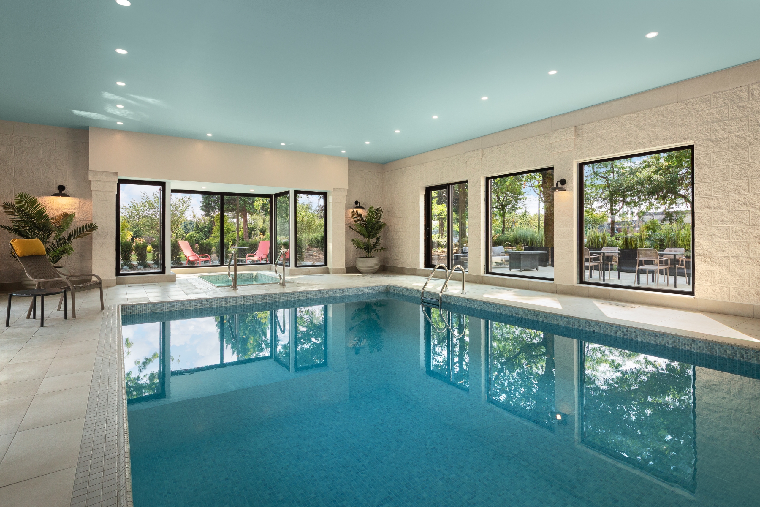 Indoor pool area with a blue ceiling, large windows, and lounge chairs by the poolside plants.