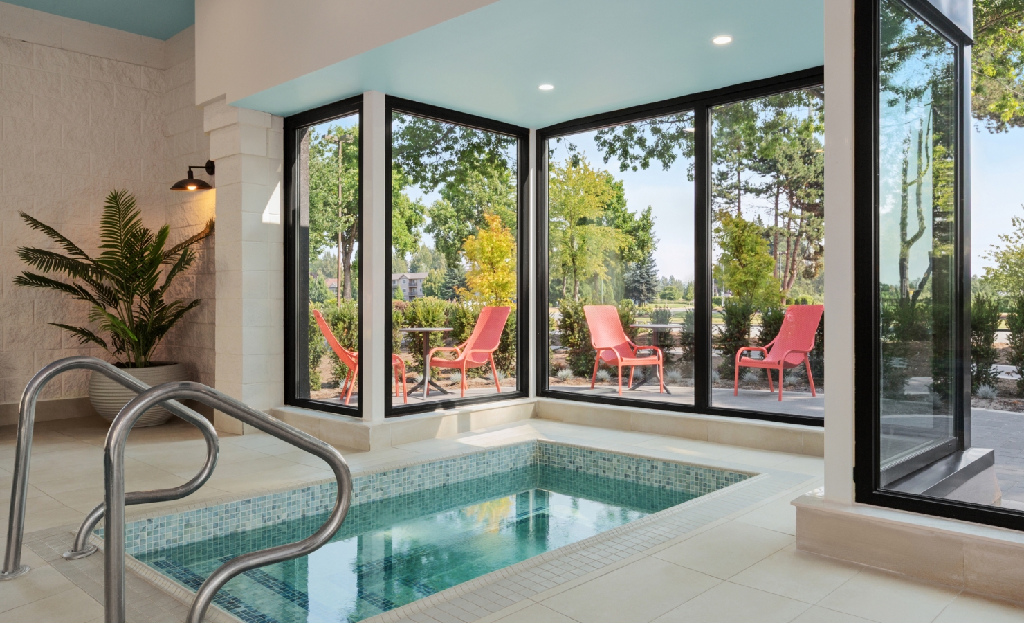 Indoor pool area with turquoise ceiling and glass walls; outdoor patio with red chairs and green trees visible outside the windows.