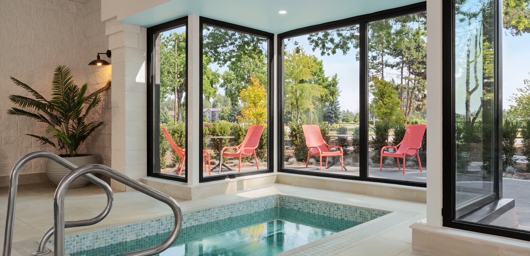 Indoor pool area with turquoise ceiling and glass walls; outdoor patio with red chairs and green trees visible outside the windows.